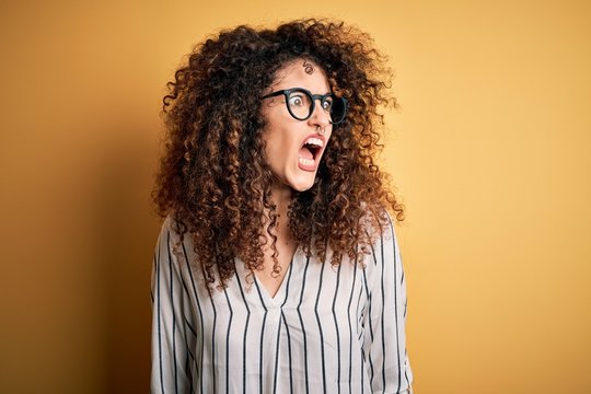 Young beautiful woman with curly hair and piercing wearing striped shirt and glasses angry and mad screaming frustrated and furious, shouting with anger. Rage and aggressive concept.