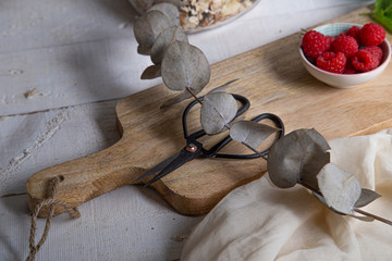 Dried leaves, wooden board, vintage scissors and a small bowl with raspberries on white table.