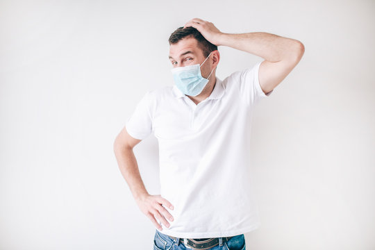 Young Sick Man Isolated Over White Background. Guy Wear Facial Medical Protection Mask To Avoid Flu And Virus. Confused Man Hold Hand On Head And Look To Left.