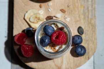 Close-up with aerial view of a glass jar with plain yogurt, blueberries, muesli and raspberries on a wooden board.