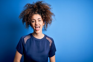Fototapeta na wymiar Young beautiful woman with curly hair and piercing wearing casual blue t-shirt sticking tongue out happy with funny expression. Emotion concept.