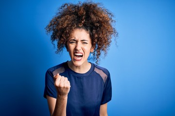 Fototapeta na wymiar Young beautiful woman with curly hair and piercing wearing casual blue t-shirt angry and mad raising fist frustrated and furious while shouting with anger. Rage and aggressive concept.