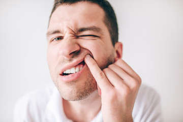 Fototapeta premium Young man isolated over white background. Close up portrait of irritated guy looking on camera and holding upper lip with one finger. Suffer from tooth pain and ache.