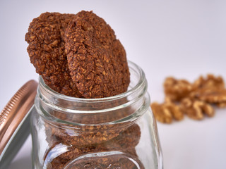 Open lid glass cookie jar with oatmeal cookies inside with walnuts in the background on white surface table - delicious gluten-free oatmeal cookies with walnuts and cocoa