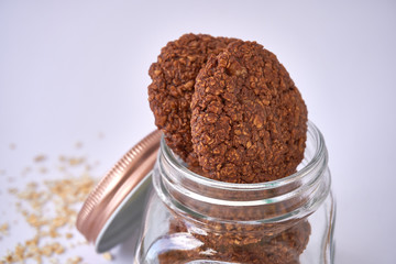 Open lid glass cookie jar with oatmeal cookies inside with oat flakes spread on the white surface table - delicious gluten-free oatmeal cookies with walnuts and cocoa
