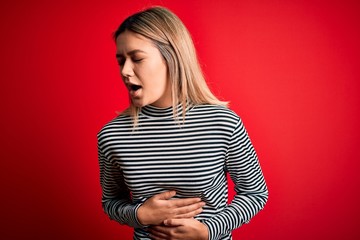 Fototapeta premium Young beautiful blonde woman wearing casual striped sweater over red isolated background with hand on stomach because nausea, painful disease feeling unwell. Ache concept.