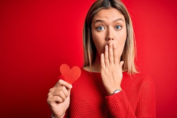 Young beautiful woman holding heart card standing over isolated red background cover mouth with hand shocked with shame for mistake, expression of fear, scared in silence, secret concept