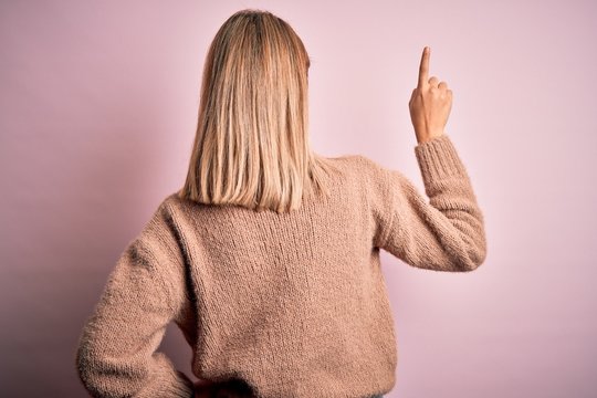 Young beautiful blonde woman wearing winter wool sweater over pink isolated background Posing backwards pointing ahead with finger hand