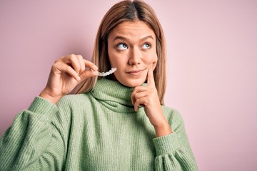 Young beautiful woman holding aligner standing over isolated pink background serious face thinking about question, very confused idea
