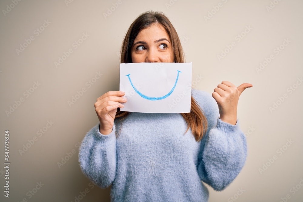 Sticker Young blonde woman holding funny smile drawing on mouth as happy expression pointing and showing with thumb up to the side with happy face smiling