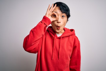 Young beautiful asian girl wearing casual sweatshirt with hoodie over white background doing ok gesture shocked with surprised face, eye looking through fingers. Unbelieving expression.
