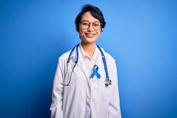 Young beautiful asian doctor girl wearing stethoscope and coat with blue cancer ribbon with a happy and cool smile on face. Lucky person.