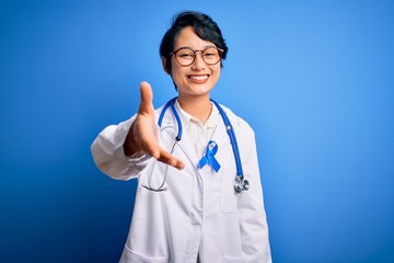 Fototapeta premium Young beautiful asian doctor girl wearing stethoscope and coat with blue cancer ribbon smiling friendly offering handshake as greeting and welcoming. Successful business.