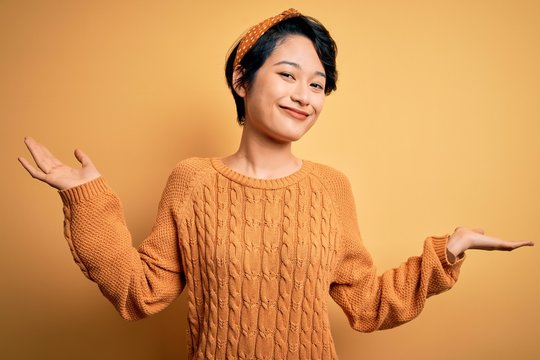 Young beautiful asian girl wearing casual sweater and diadem standing over yellow background clueless and confused expression with arms and hands raised. Doubt concept.