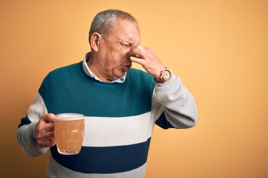 Senior Handsome Man Drinking Jar Of Beer Standing Over Isolated Yellow Background Smelling Something Stinky And Disgusting, Intolerable Smell, Holding Breath With Fingers On Nose. Bad Smell