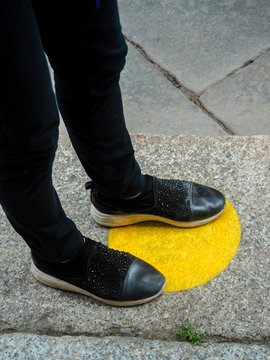 Human Legs And Feet Waiting In Line And Standing Inside Marked Yellow Circles With Social Distancing Measures. Yellow Road Marking And Sidewalk Symbol.