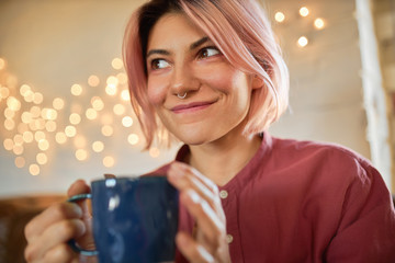 Close up shot of charming 20 year old female with facial piercing and pink hair looking up with mysterious smile, drinking coffee from mug, enjoying nice aroma, posing against blurred light background
