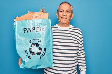 Senior grey-haired man recycling holding bag with cardboard to recycle over blue background with a happy face standing and smiling with a confident smile showing teeth