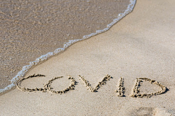 Covid written on the sand of a beach with wave washing, erasing or canceling it during Coronavirus summer, Covid free, safe beach
