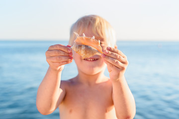 Blond boy holding seashell and smiling. Vacation with children of the sea. Happy childhood concept