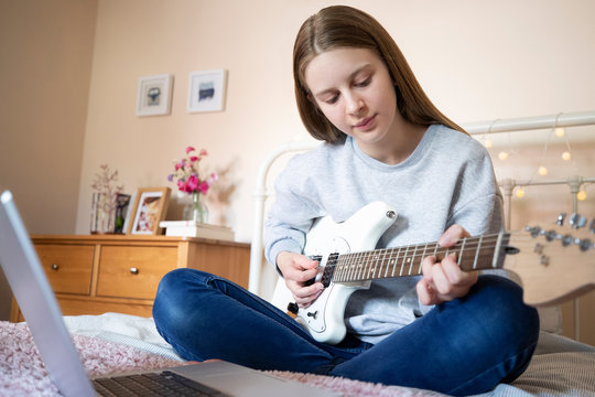Teenage Girl Sitting On Bed Learning To Play Electric Guitar With Online Lesson On Laptop Computer