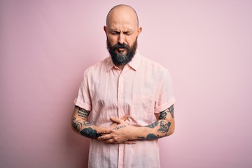 Handsome bald man with beard and tattoo wearing casual shirt over isolated pink background with hand on stomach because indigestion, painful illness feeling unwell. Ache concept.