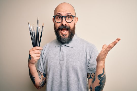 Handsome bald artist man with beard and tattoo painting using painter brushes very happy and excited, winner expression celebrating victory screaming with big smile and raised hands