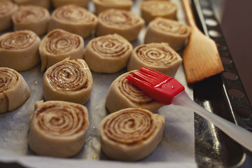 cooking homemade cinnabon buns from yeast dough on black aking sheet with baking paper and red silicone brush