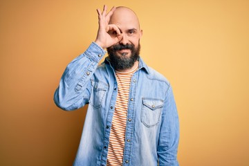 Handsome bald man with beard wearing casual denim jacket and striped t-shirt doing ok gesture with hand smiling, eye looking through fingers with happy face.