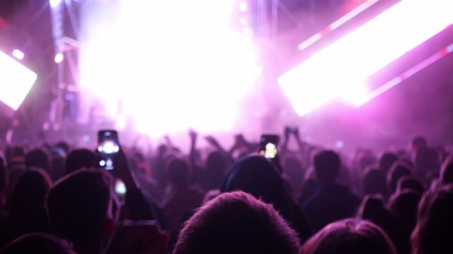 People make photos and videos at the festival. The crowd watches a concert, sings, jumps. Strobing stage lights. Happy people are watching an amazing concert. World tour of popular musicians.