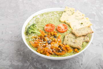 Green broccoli cream soup with crackers in white bowl on a gray concrete background, side view.