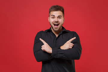 Surprised young bearded guy 20s in classic black shirt posing isolated on red background studio portrait. People lifestyle concept. Mock up copy space. Pointing index fingers aside hold hands crossed.