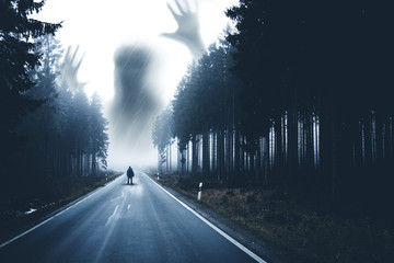 Mann steht auf einer Straße im dunklen Wald. Geisterhafte Figur taucht im Himmel auf. - obrazy, fototapety, plakaty