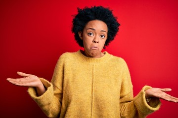 Fototapeta na wymiar Young beautiful African American afro woman with curly hair wearing casual yellow sweater clueless and confused expression with arms and hands raised. Doubt concept.