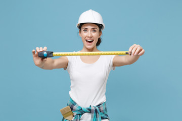 Cheerful young woman in casual clothes protective helmet hardhat hold measure tape isolated on blue background in studio. Instruments accessories for renovation apartment room. Repair home concept.