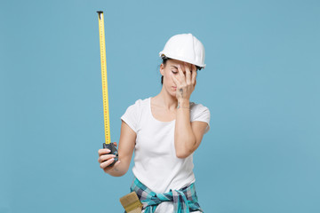 Exhausted young woman in protective helmet hardhat hold measure tape isolated on blue background studio. Instruments accessories for renovation apartment room. Repair home concept. Put hand on face.