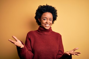 Young beautiful African American afro woman with curly hair wearing casual turtleneck sweater smiling cheerful with open arms as friendly welcome, positive and confident greetings