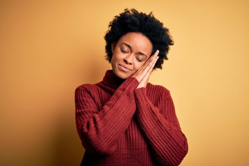 Young beautiful African American afro woman with curly hair wearing casual turtleneck sweater sleeping tired dreaming and posing with hands together while smiling with closed eyes.