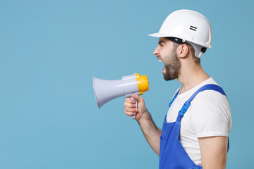 Side view of crazy young man in coveralls protective helmet hardhat scream in megaphone isolated on blue background studio. Instruments accessories for renovation apartment room. Repair home concept.