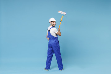 Back rear view of cheerful young man in coveralls protective helmet hardhat hold paint roller isolated on blue background. Instruments accessories for renovation apartment room. Repair home concept.