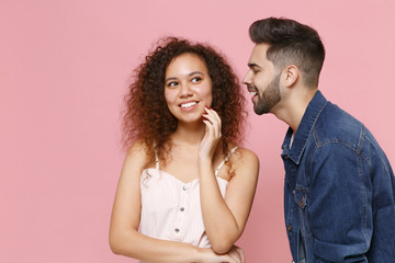 Cheerful young couple friends european guy african american girl in casual clothes isolated on pastel pink background in studio. People lifestyle concept. Whispering secret, sharing news near ear.