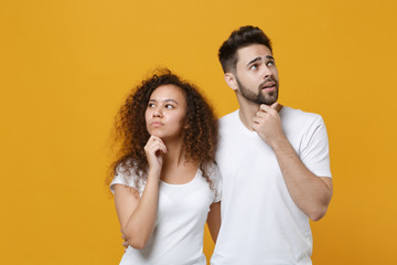Pensive young couple two friends european guy african american girl in white t-shirts posing isolated on yellow wall background. People lifestyle concept. Put hands prop up on chin, looking aside up.