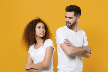 Confused young couple friends european guy african american girl in white t-shirts isolated on yellow background. People lifestyle concept. Stand back to back looking at each other hold hands crossed.