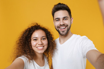 Obraz premium Close up of smiling couple friends european guy african american girl in white t-shirts isolated on yellow background. People lifestyle concept. Mock up copy space. Doing selfie shot on mobile phone.