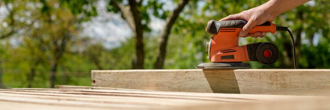 Unrecognizable man in the garden sanding wooden planks. DIY home improvement, restoration, carpentry concept. Hand detail banner with copy space.
