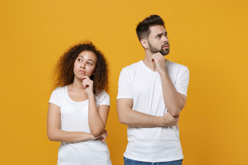 Confused pensive young couple two friends european guy african american girl in white t-shirts posing isolated on yellow background. People lifestyle concept. Put hand prop up on chin, looking aside.