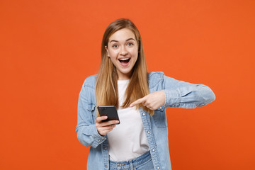 Excited young woman girl in casual denim clothes posing isolated on bright orange wall background studio portrait. People lifestyle concept. Mock up copy space. Pointing index finger on mobile phone.