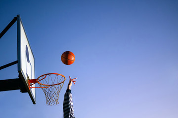 Street basketball ball player throwing ball into the hoop. Close up of hand, orange ball above the hoop net with blue sky in the background. Concept of success, scoring points and winning. Copy space © CrispyMedia