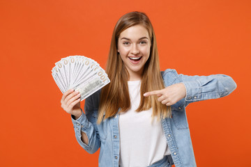 Excited young woman girl in casual denim clothes posing isolated on orange background. People lifestyle concept. Mock up copy space. Pointing index finger on fan of cash money in dollar banknotes.