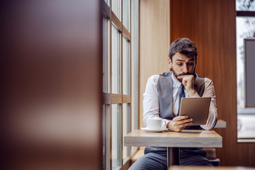 Bearded businessman sitting in coffee shop and reading news on tablet.